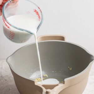 A hand pours milk from a measuring cup into a saucepan containing small food pieces, on a textured surface.