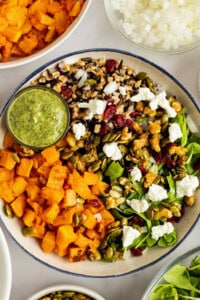 A salad with mixed greens, roasted squash, goat cheese, nuts, seeds, and grains, served with a small bowl of green dressing on the side.