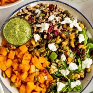 A salad with mixed greens, roasted squash, goat cheese, nuts, seeds, and grains, served with a small bowl of green dressing on the side.