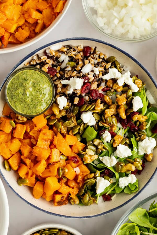 A salad with mixed greens, roasted squash, goat cheese, nuts, seeds, and grains, served with a small bowl of green dressing on the side.