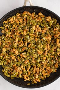 A pan filled with a mix of nuts and seeds, predominantly featuring green pumpkin seeds and walnuts, with a light coating, likely for roasting.