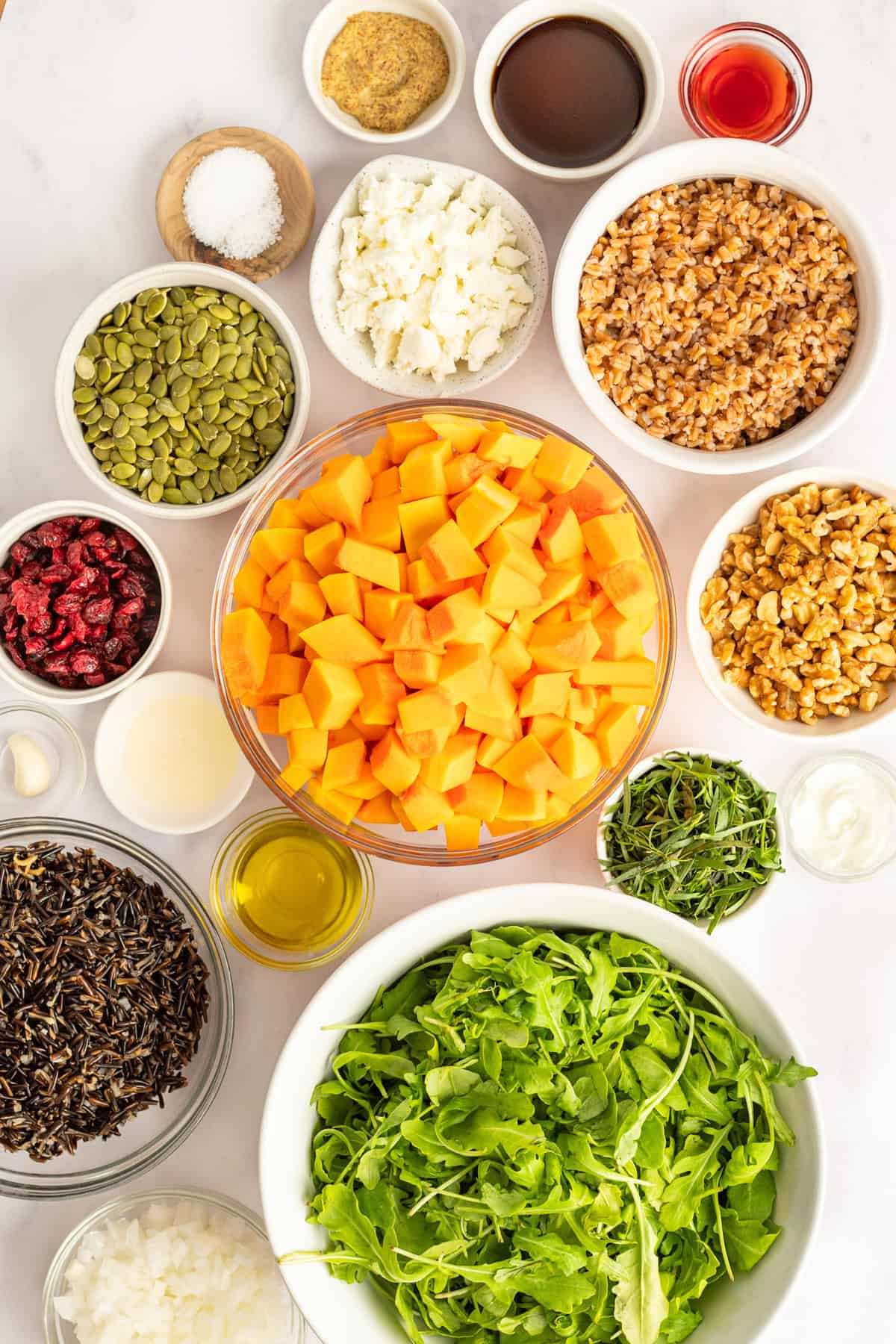 Ingredients on a table including arugula, cubed squash, nuts, seeds, grains, cheese, and various liquids in bowls, viewed from above.