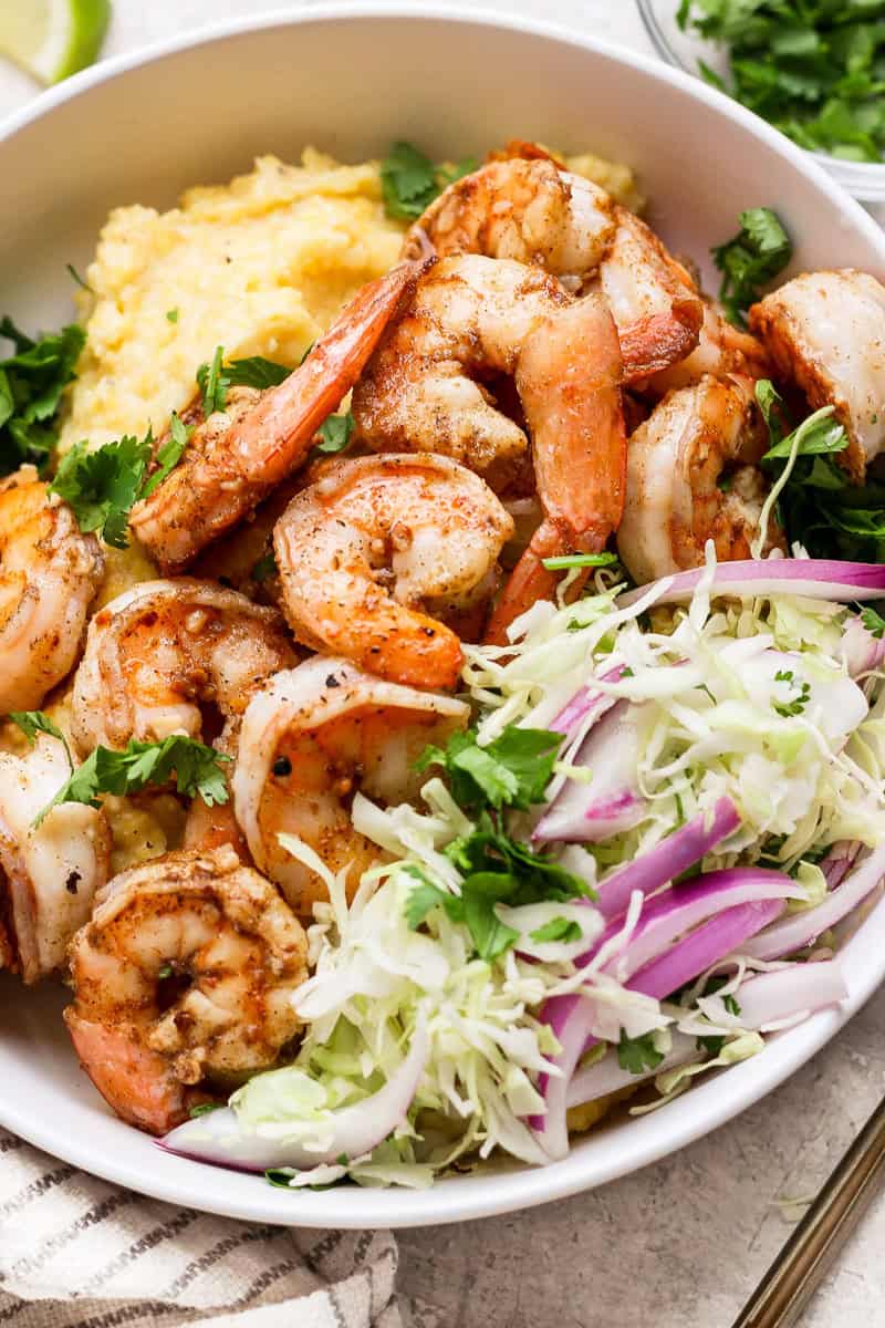 A bowl of seasoned shrimp atop creamy polenta, garnished with fresh herbs, sliced cabbage, red onion, and cilantro. A lime wedge is partially visible on the side.