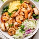 A bowl of seasoned shrimp with creamy grits, garnished with fresh cilantro, alongside a cabbage and red onion salad.