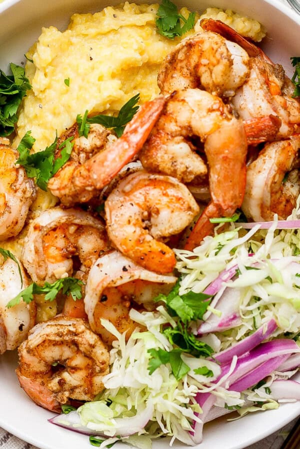 A bowl of seasoned shrimp with creamy grits, garnished with fresh cilantro, alongside a cabbage and red onion salad.