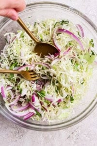 A hand mixing coleslaw in a clear glass bowl with a fork and spoon. The coleslaw contains shredded cabbage, red onions, and herbs.