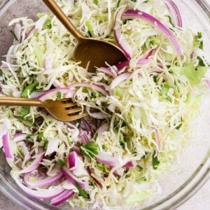 A hand mixing coleslaw in a clear glass bowl with a fork and spoon. The coleslaw contains shredded cabbage, red onions, and herbs.