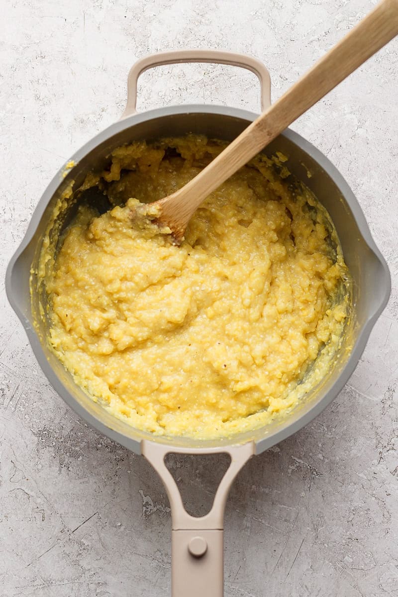 A pot on a light surface contains thick yellow grits being stirred with a wooden spoon.