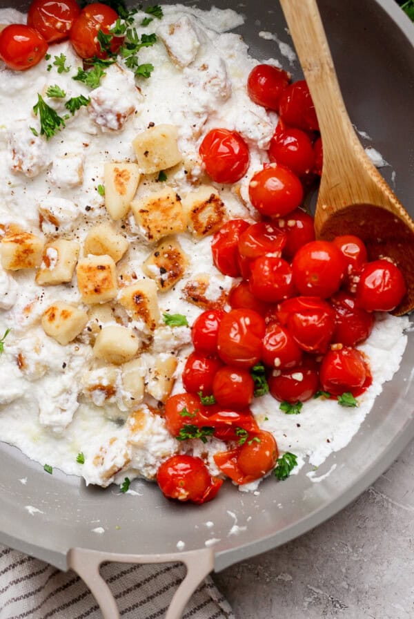 Pan with cooked cherry tomatoes, gnocchi, and white sauce, garnished with herbs, and a wooden spoon on the side.