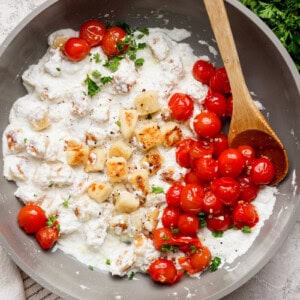 A skillet with melted cheese, roasted cherry tomatoes, and herbs, accompanied by a wooden spoon. Fresh parsley is on the side.