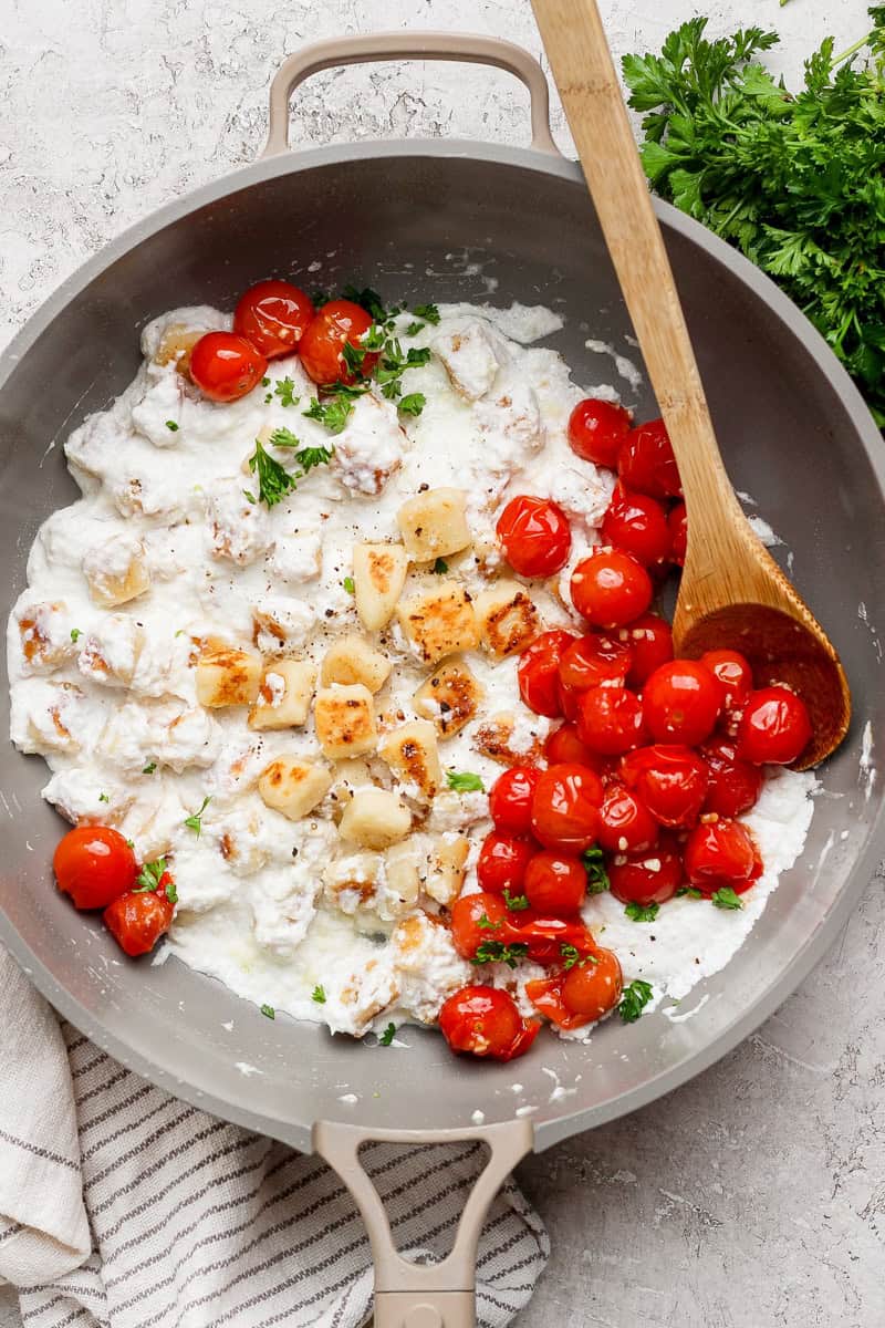 A skillet with melted cheese, roasted cherry tomatoes, and herbs, accompanied by a wooden spoon. Fresh parsley is on the side.