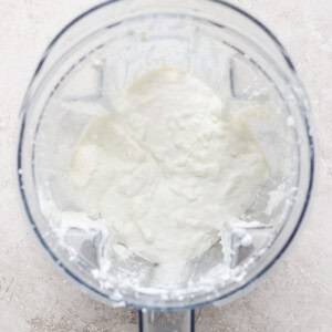 A blender container with a creamy white mixture inside, viewed from above, on a light textured surface.