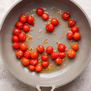 Cherry tomatoes and minced garlic are sautéing in olive oil in a beige frying pan placed on a light textured surface.