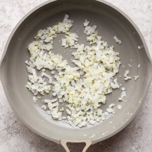 Chopped onions and garlic sautéing in a large frying pan on a light textured surface.