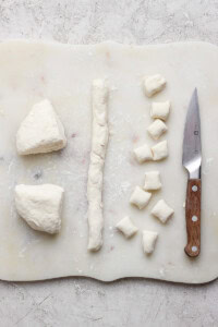 Dough is cut into small pieces on a white cutting board with a wooden-handled knife beside them.