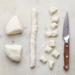 Dough is cut into small pieces on a white cutting board with a wooden-handled knife beside them.