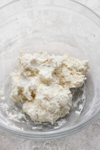 A clear bowl containing a mixture of flour and butter, forming a crumbly dough.