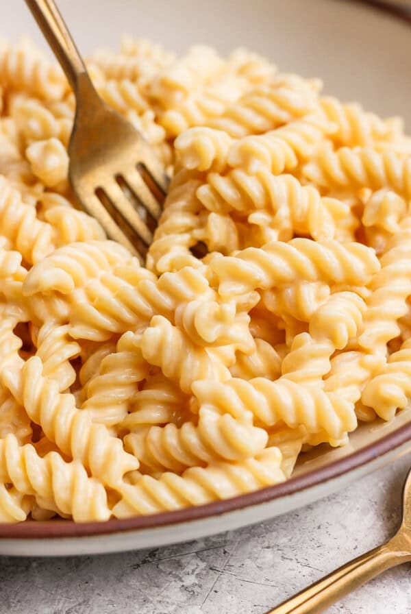 A fork twirls creamy cheese sauce-covered rotini pasta in a bowl on a textured surface.