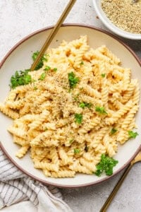 A bowl of rotini pasta with creamy sauce, topped with chopped herbs and breadcrumbs, on a round plate with a fork.