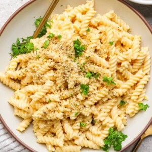 A bowl of rotini pasta with creamy sauce, topped with chopped herbs and breadcrumbs, on a round plate with a fork.