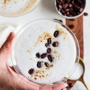 A hand holding a frothy coffee drink in a glass, garnished with coffee beans and cinnamon. Another glass and a bowl of coffee beans are visible in the background.