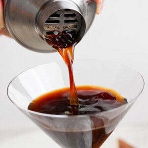 A hand pouring a dark liquid from a metal shaker into a martini glass.