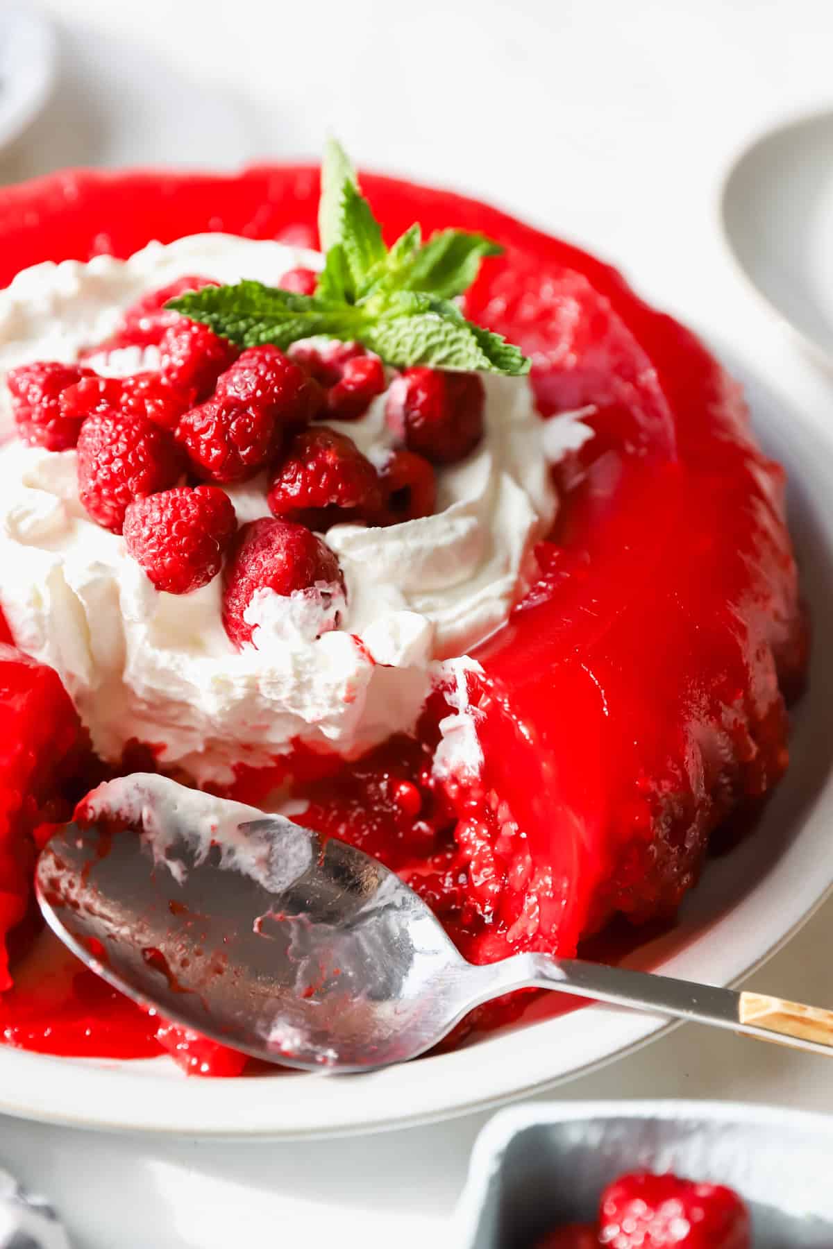 A red gelatin dessert topped with whipped cream, fresh raspberries, and a mint sprig. A spoon rests on the plate beside it.