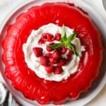 A red gelatin dessert topped with whipped cream, fresh raspberries, and mint leaves on a white plate.