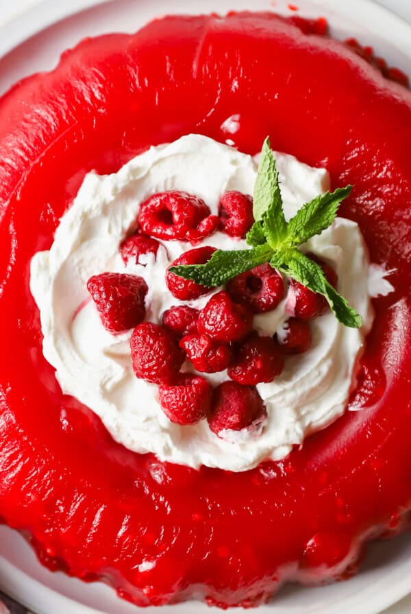 A red gelatin dessert topped with whipped cream, fresh raspberries, and mint leaves on a white plate.