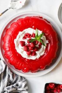 A red gelatin dessert with whipped cream, raspberries, and mint leaves on top, served on a white plate. A striped cloth and small dish of raspberries are nearby.