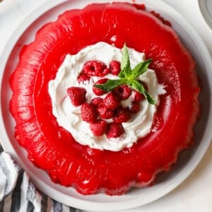 A red gelatin dessert with whipped cream, raspberries, and mint leaves on top, served on a white plate. A striped cloth and small dish of raspberries are nearby.