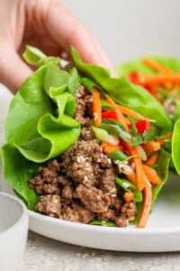 A hand holding a lettuce wrap filled with ground meat, shredded carrots, and herbs on a white plate.