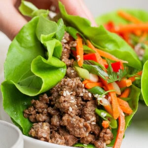 A hand holding a lettuce wrap filled with ground meat, shredded carrots, and herbs on a white plate.