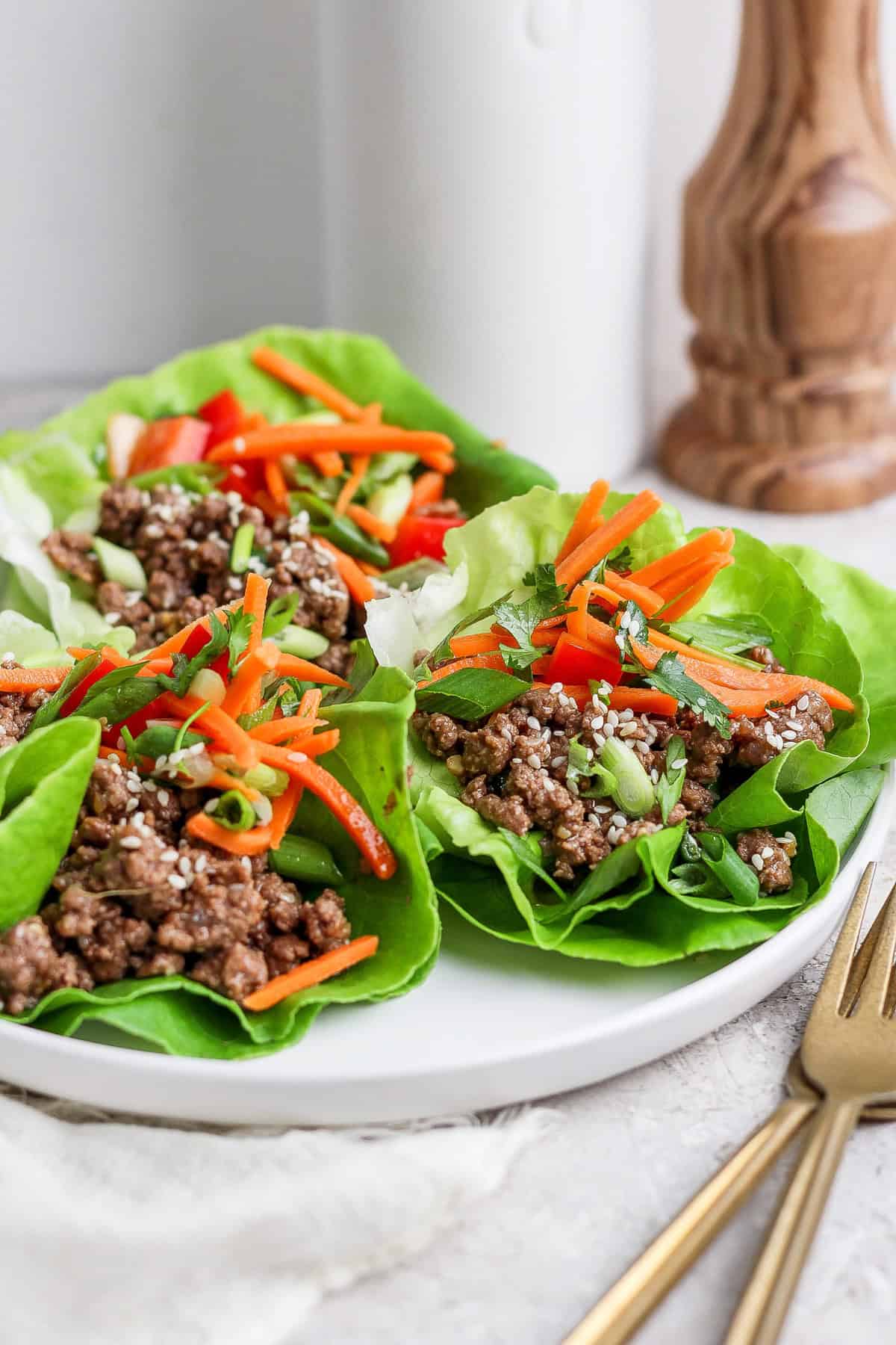 Lettuce wraps filled with minced meat, sliced carrots, sesame seeds, and green onions on a white plate. Gold cutlery is visible in the foreground.