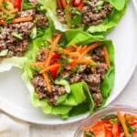 Lettuce wraps on a plate, filled with seasoned ground beef, sliced carrots, green onions, and sesame seeds, with a side bowl of carrots and red peppers.