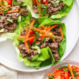 Lettuce wraps on a plate, filled with seasoned ground beef, sliced carrots, green onions, and sesame seeds, with a side bowl of carrots and red peppers.