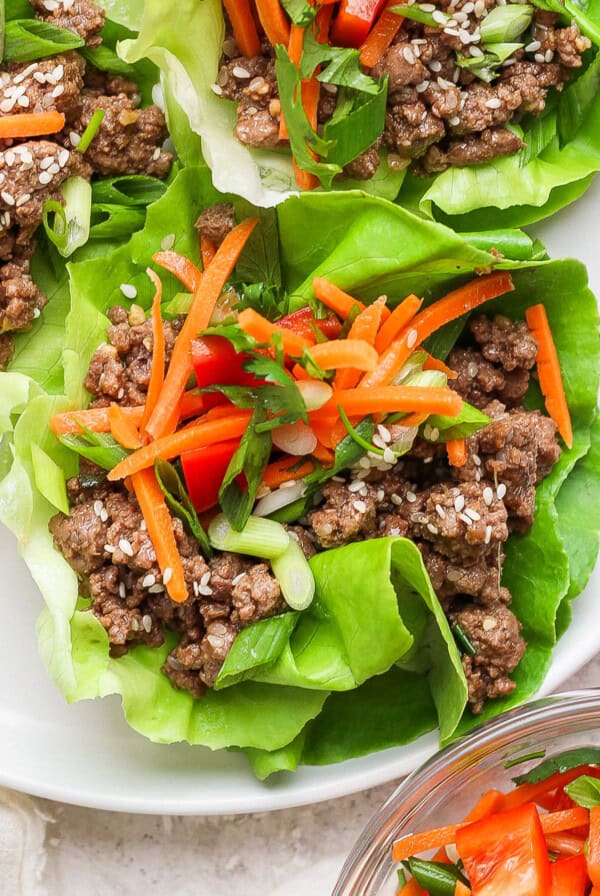 Lettuce wraps on a plate, filled with seasoned ground beef, sliced carrots, green onions, and sesame seeds, with a side bowl of carrots and red peppers.