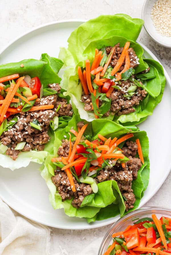 Four lettuce wraps filled with ground meat, shredded carrots, diced red bell peppers, and green onions, served on a white plate.