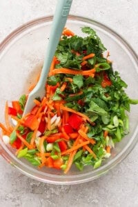 A glass bowl filled with chopped vegetables, including red bell peppers, carrots, green onions, cilantro, and green bell peppers, with a light blue spatula.