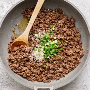 Ground beef cooked in a pan with a wooden spoon, topped with sesame seeds and chopped green onions.