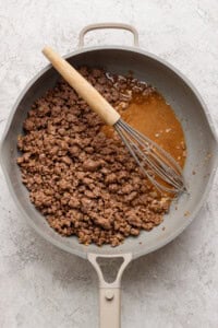 Ground beef cooking in a skillet with a whisk resting on the side and a pool of juices visible.