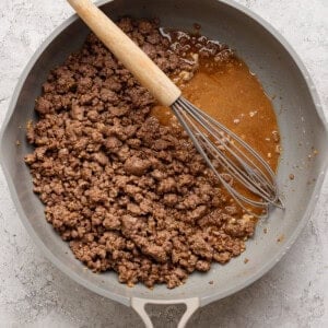 Ground beef cooking in a skillet with a whisk resting on the side and a pool of juices visible.