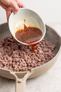 A hand pours a brown sauce from a white bowl onto cooked ground beef in a beige skillet.