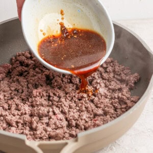 A hand pours a brown sauce from a white bowl onto cooked ground beef in a beige skillet.