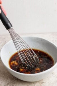 A whisk is stirring a dark-colored sauce in a light gray bowl on a textured surface.