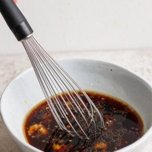 A whisk is stirring a dark-colored sauce in a light gray bowl on a textured surface.