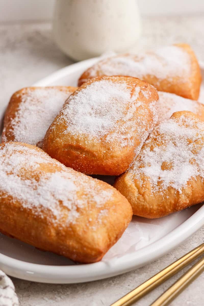 A plate of rectangular beignets coated with powdered sugar on a light-colored surface.