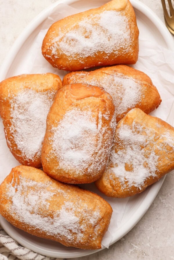 Six sugar-coated beignets stacked on a white oval plate.