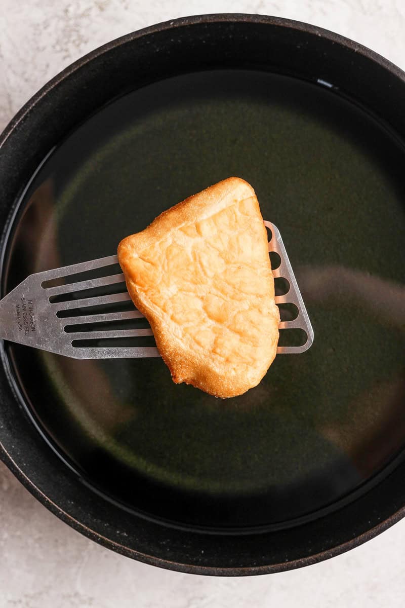 A piece of fried bread being lifted from a pan of oil with a metal spatula.