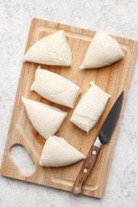 Six pieces of dough are placed on a wooden cutting board next to a knife.